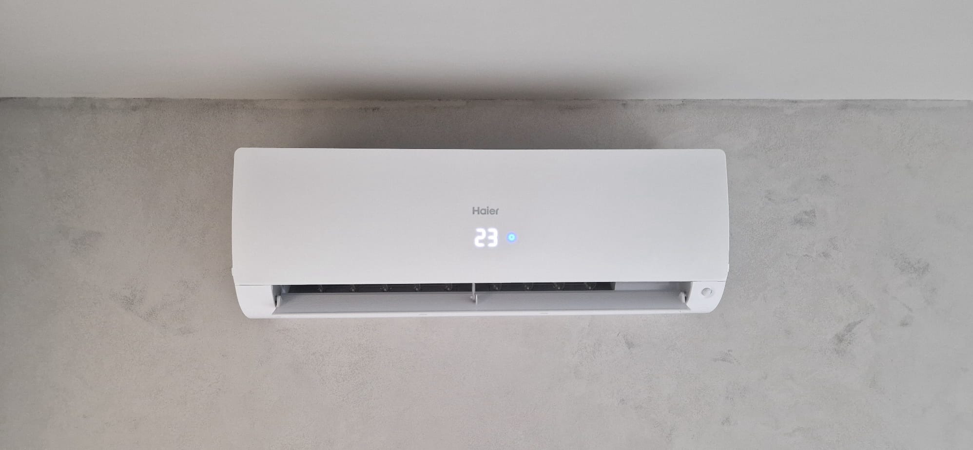 Biała klimatyzacja Haier na szarej ścianie z efektem betonu. Widoczny wyświetlacz z temperaturą 23 stopnie. Minimalistyczny design, nowoczesne wnętrze.