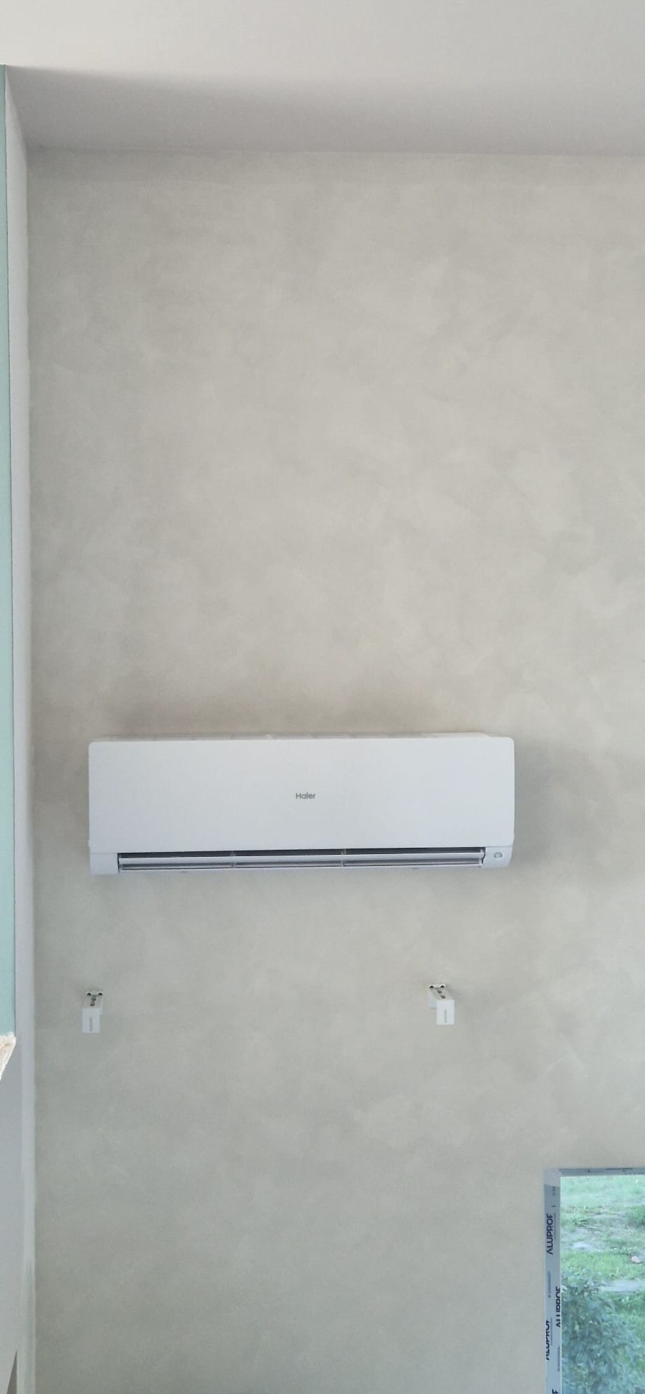 Biały klimatyzator Haier zamontowany na ścianie o fakturze betonu, widoczne uchwyty montażowe i fragment okna z profilem Aluplast. Ujęcie z dołu, neutralne oświetlenie.