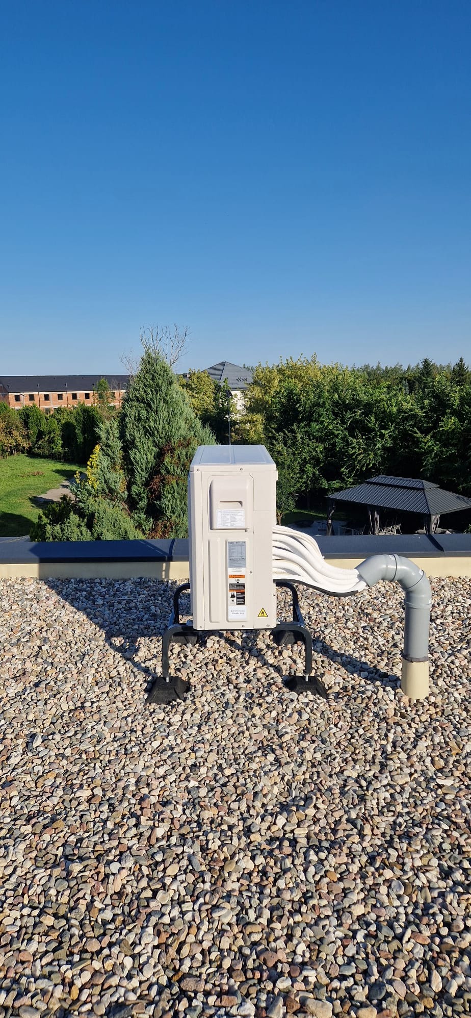 Biała jednostka zewnętrzna klimatyzacji na dachu pokrytym kamieniami, z widocznymi przewodami i rurą odprowadzającą skropliny, na tle zieleni i błękitnego nieba.