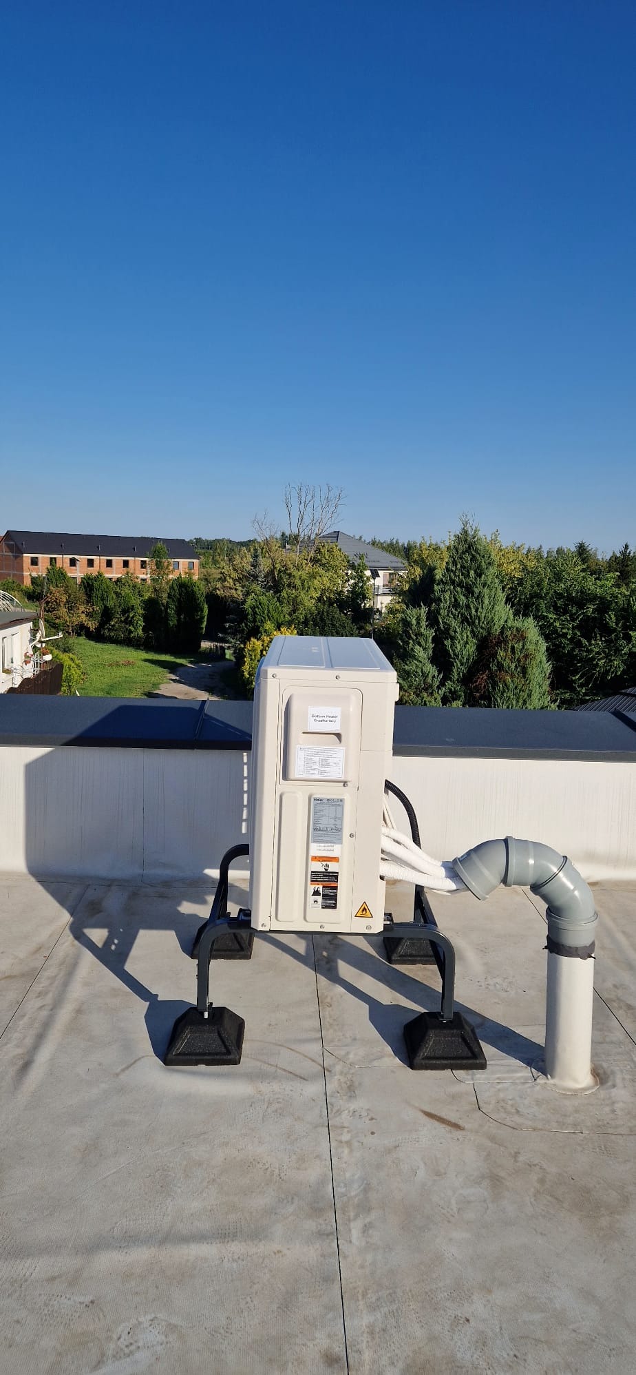 Biała jednostka zewnętrzna klimatyzacji na dachu budynku, z widocznymi przewodami i elementami montażowymi. W tle zieleń i budynki mieszkalne.