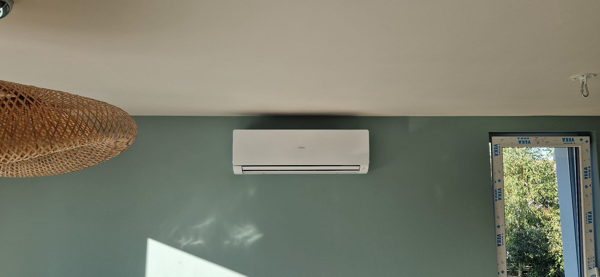 Biała klimatyzacja Haier na zielonej ścianie w pokoju z oknem i plecioną lampą. Widoczny fragment sufitu i hak do lampy.