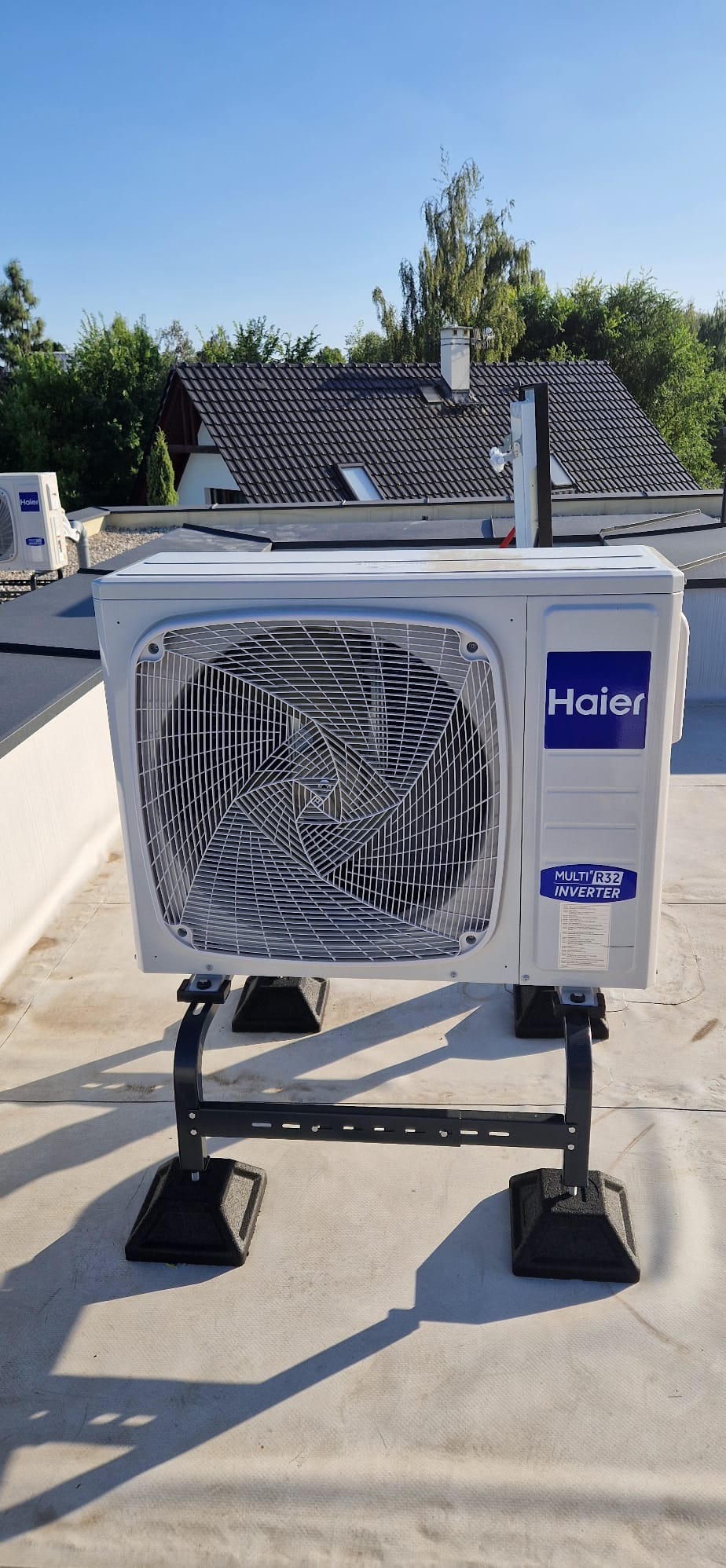 Biała jednostka zewnętrzna klimatyzacji Haier na dachu, zamontowana na czarnym stelażu. W tle dom z ciemnym dachem i zieleń drzew pod błękitnym niebem.