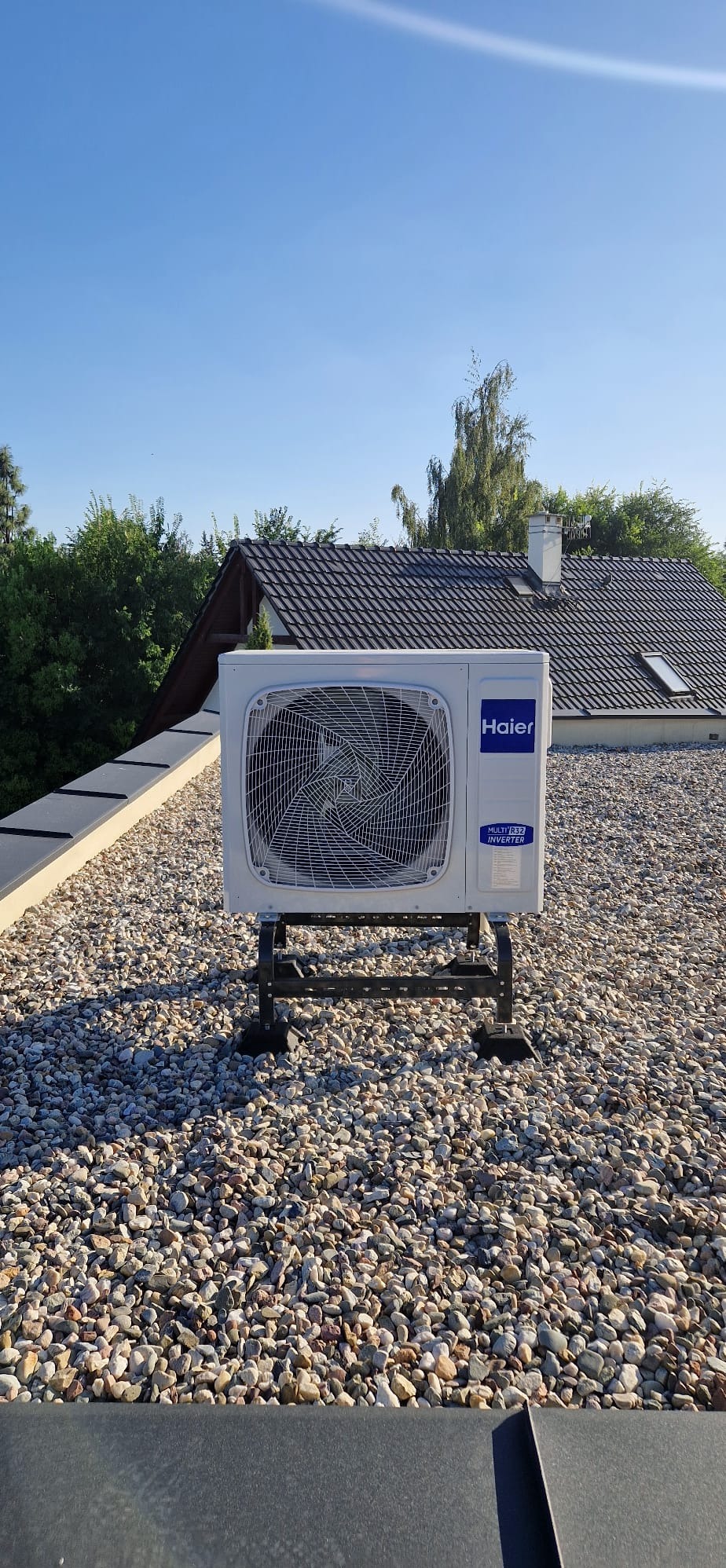 Biała jednostka zewnętrzna klimatyzacji Haier na dachu pokrytym żwirem, na metalowym stelażu. W tle budynek z ciemną dachówką i zieleń drzew.