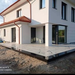 ASZTAR JAN KAJPUST - Nowoczesny dom jednorodzinny z szarym tarasem wyłożonym płytami, czarna obwódka, widoczne pręty zbrojeniowe, teren wokół w trakcie prac ziemnych.