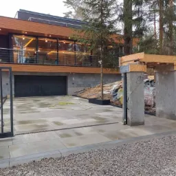 Nowoczesny podjazd z jasnoszarej kostki brukowej, z widocznymi liniowymi odwodnieniami, prowadzący do domu z garażem i drewnianą elewacją, otoczony zielenią i elementami ogrodzenia w trakcie montażu.