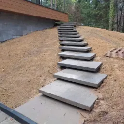 Stopnie z szarego kamienia prowadzące pod górę do nowoczesnego domu z drewnianą elewacją, otoczone piaszczystym podłożem i drzewami.