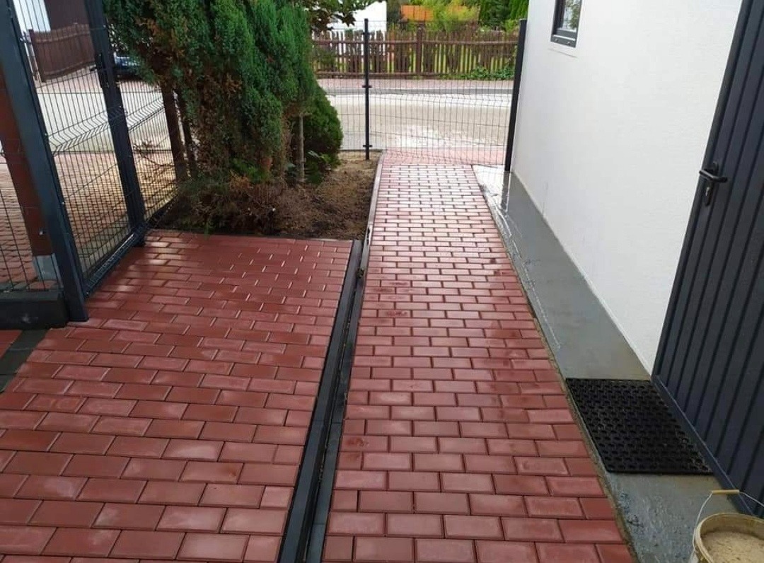 Czerwona kostka brukowa ułożona jako ścieżka wzdłuż budynku i ogrodzenia, z widocznym odpływem liniowym i zielenią w tle.