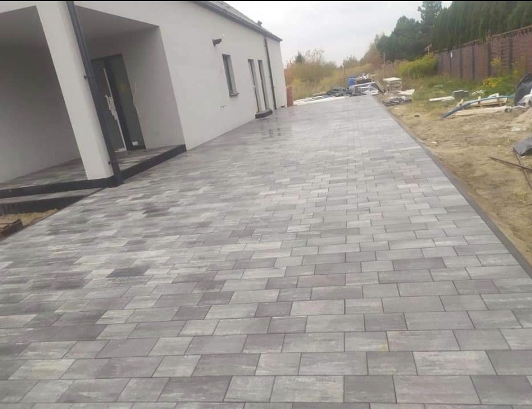 Ułożona szara kostka brukowa przed nowoczesnym domem z białymi ścianami, widoczny fragment ogrodu i ogrodzenia w tle.