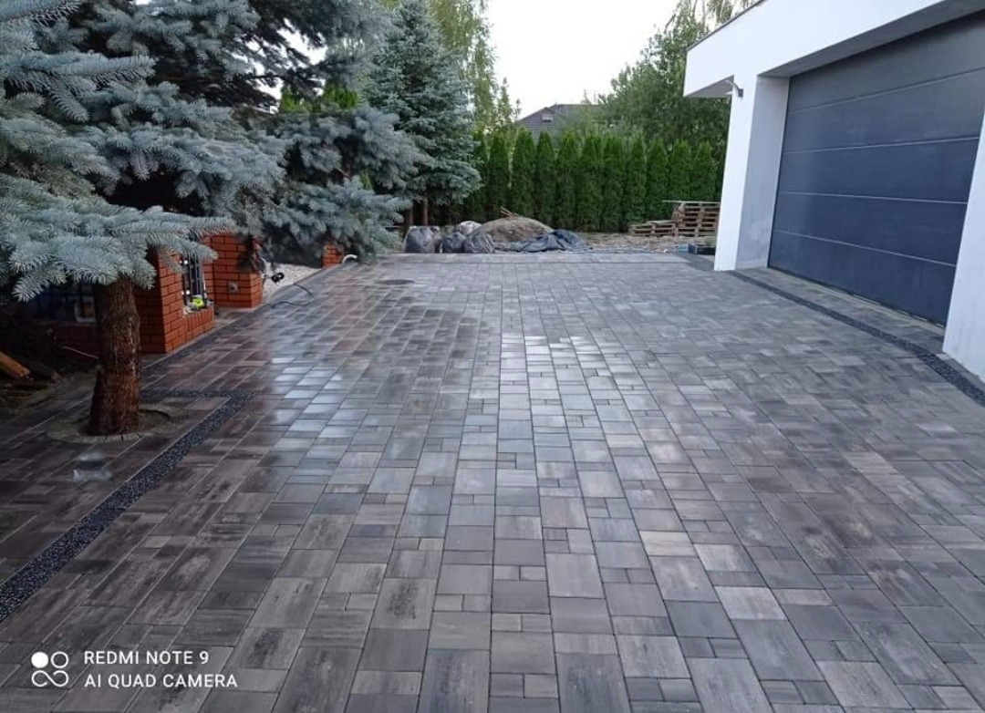 Ułożona kostka brukowa w Piasecznie na podjeździe do garażu, z widocznym fragmentem domu i zielenią w tle. Kostka w odcieniach szarości, ułożona w regularny wzór, obrzeża wykończone drobnym grysem.