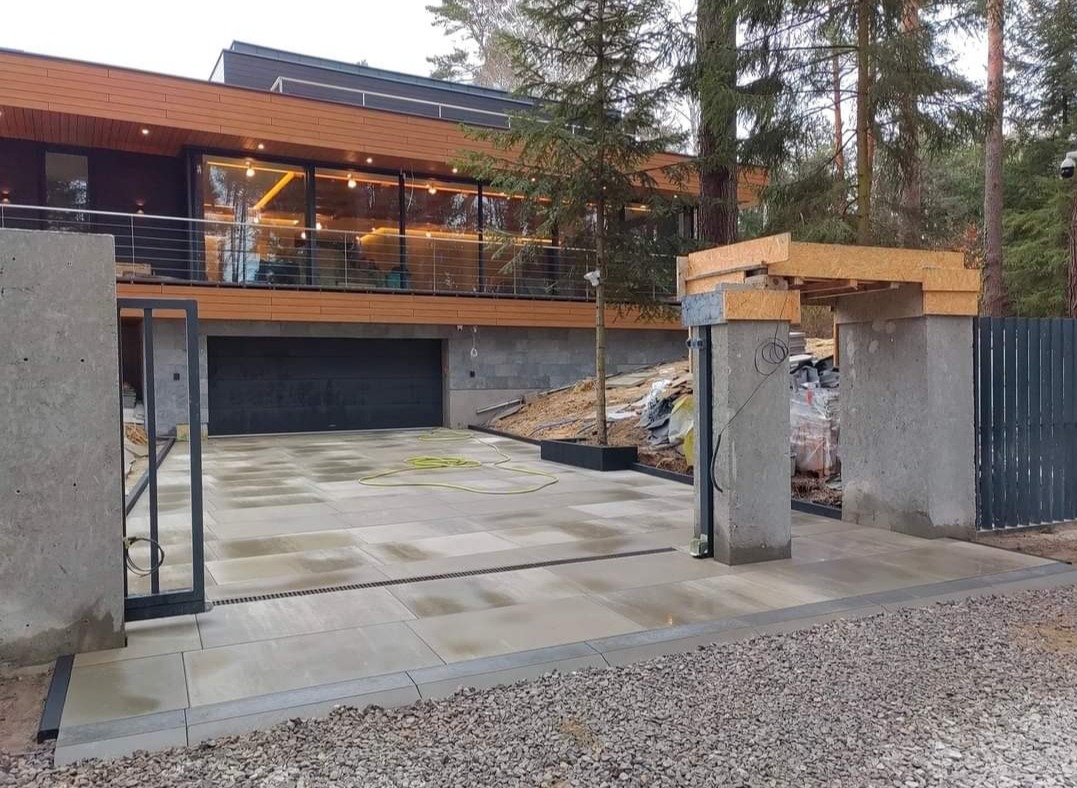 Nowoczesny podjazd z jasnoszarej kostki brukowej, z widocznymi liniowymi odwodnieniami, prowadzący do domu z garażem i drewnianą elewacją, otoczony zielenią i elementami ogrodzenia w trakcie montażu.