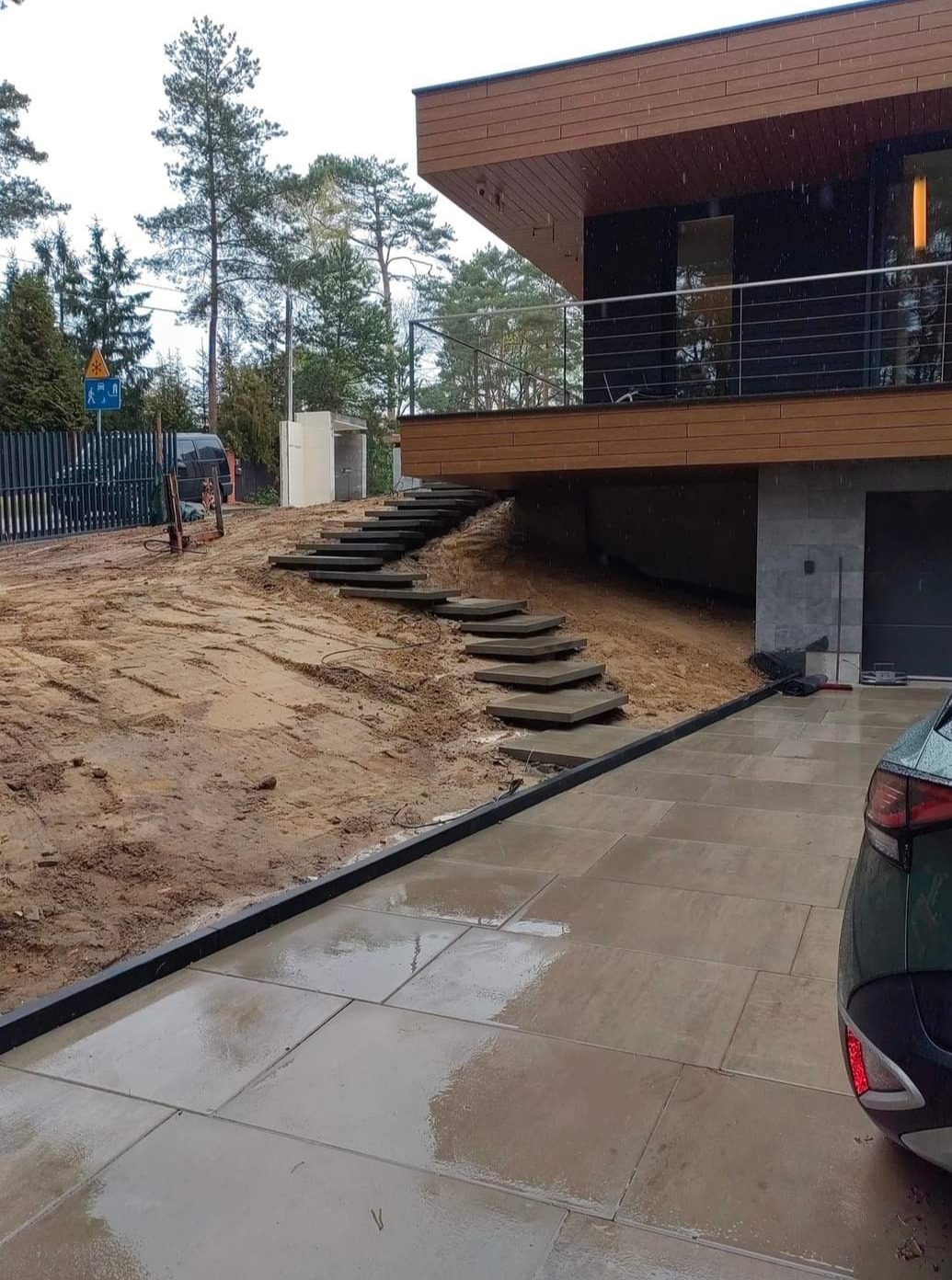Nowoczesny dom z tarasem i schodami z płyt kamiennych w trakcie budowy, widoczna barierka tarasu, elementy drenażu oraz częściowo ułożona nawierzchnia z płyt.