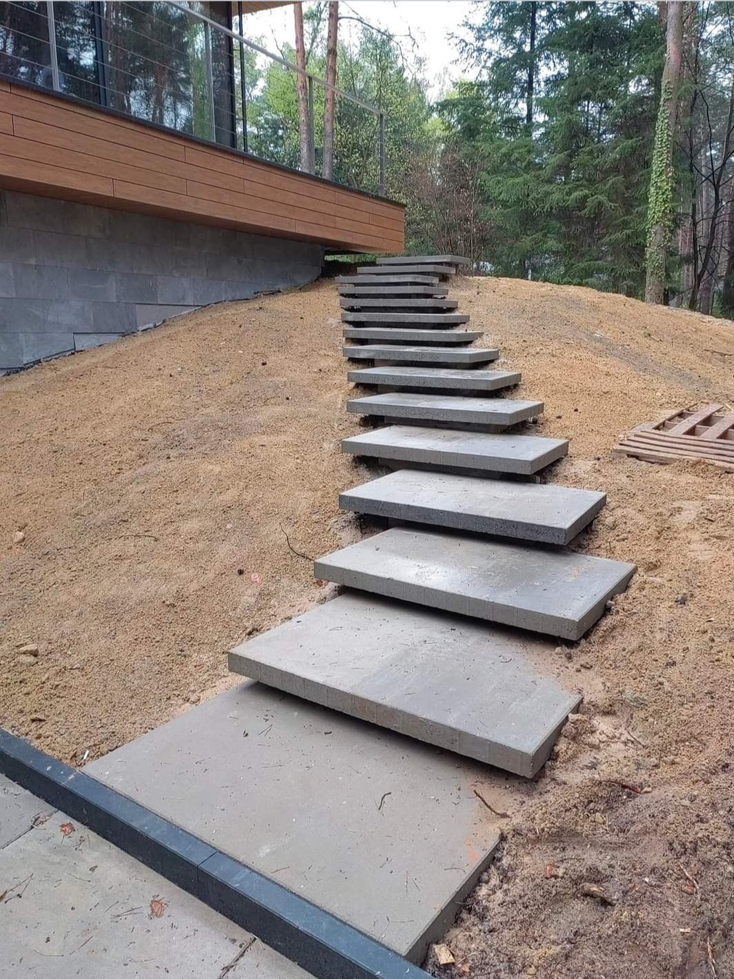 Stopnie z szarego kamienia prowadzące pod górę do nowoczesnego domu z drewnianą elewacją, otoczone piaszczystym podłożem i drzewami.