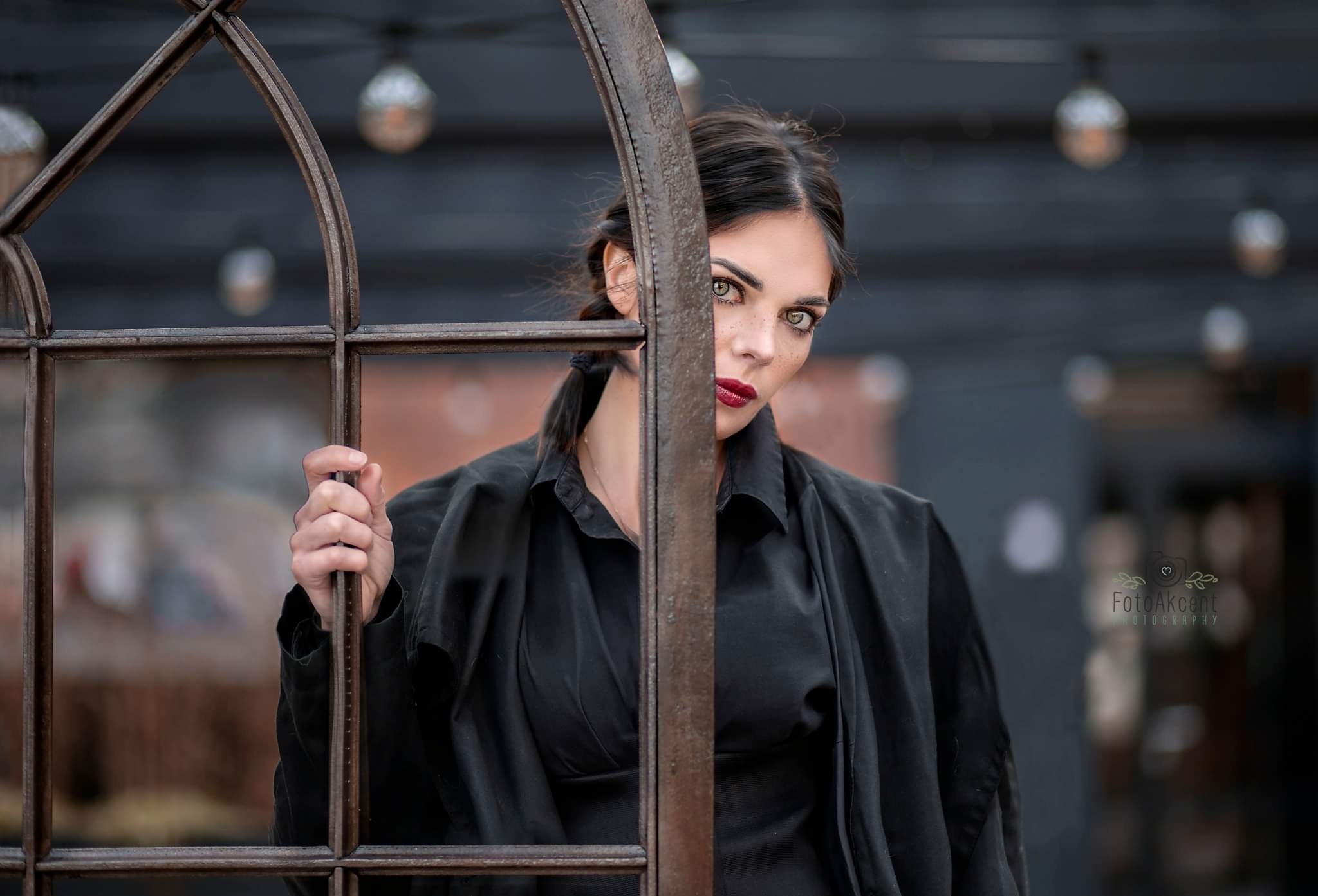 Portret kobiety o przenikliwym spojrzeniu, z rudą szminką i piegami, pozującej za metalową ramą okienną w stylu industrialnym, ubrana w czarną koszulę i płaszcz.