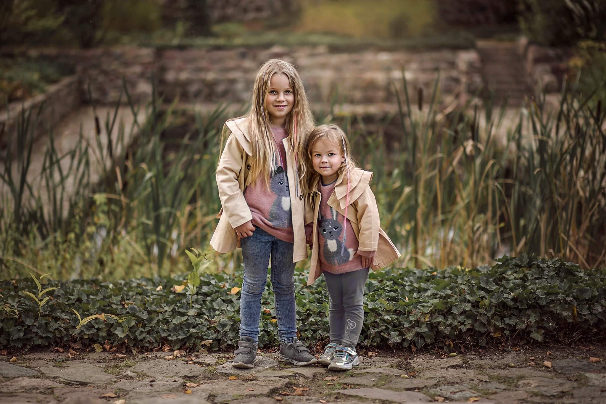 Dwie dziewczynki, blondynki, w beżowych płaszczach i swetrach z motywem królika, stojące na kamiennej ścieżce przed stawem porośniętym trzciną i bluszczem.