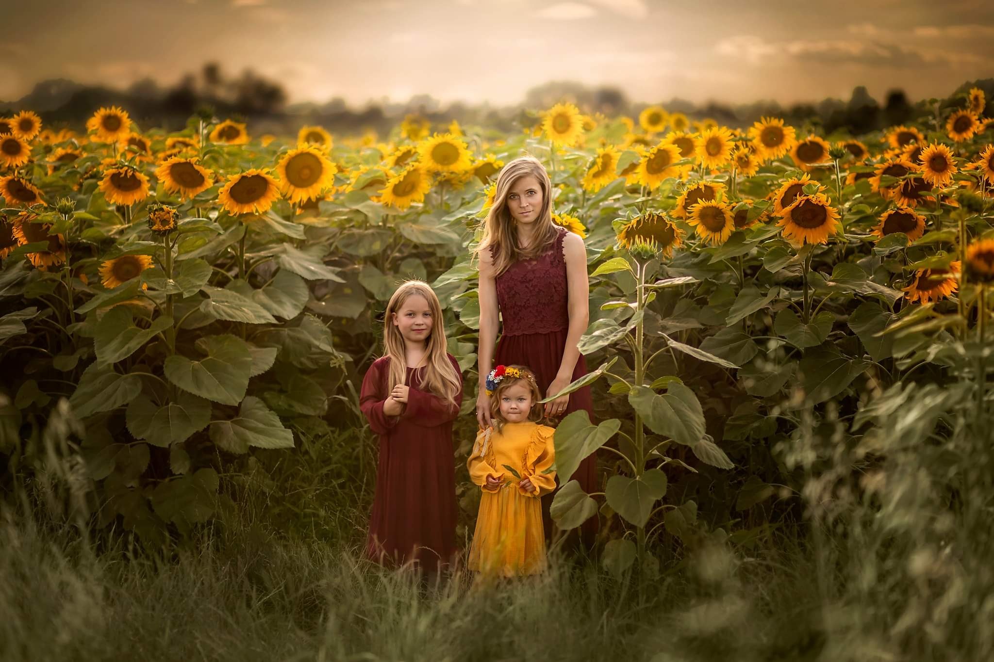 Portret matki z dwiema córkami na tle pola słoneczników w Sochaczewie, dziewczynki ubrane w sukienki, starsza w bordowej, młodsza w żółtej z wiankiem na głowie, ciepłe światło zachodzącego słońca.