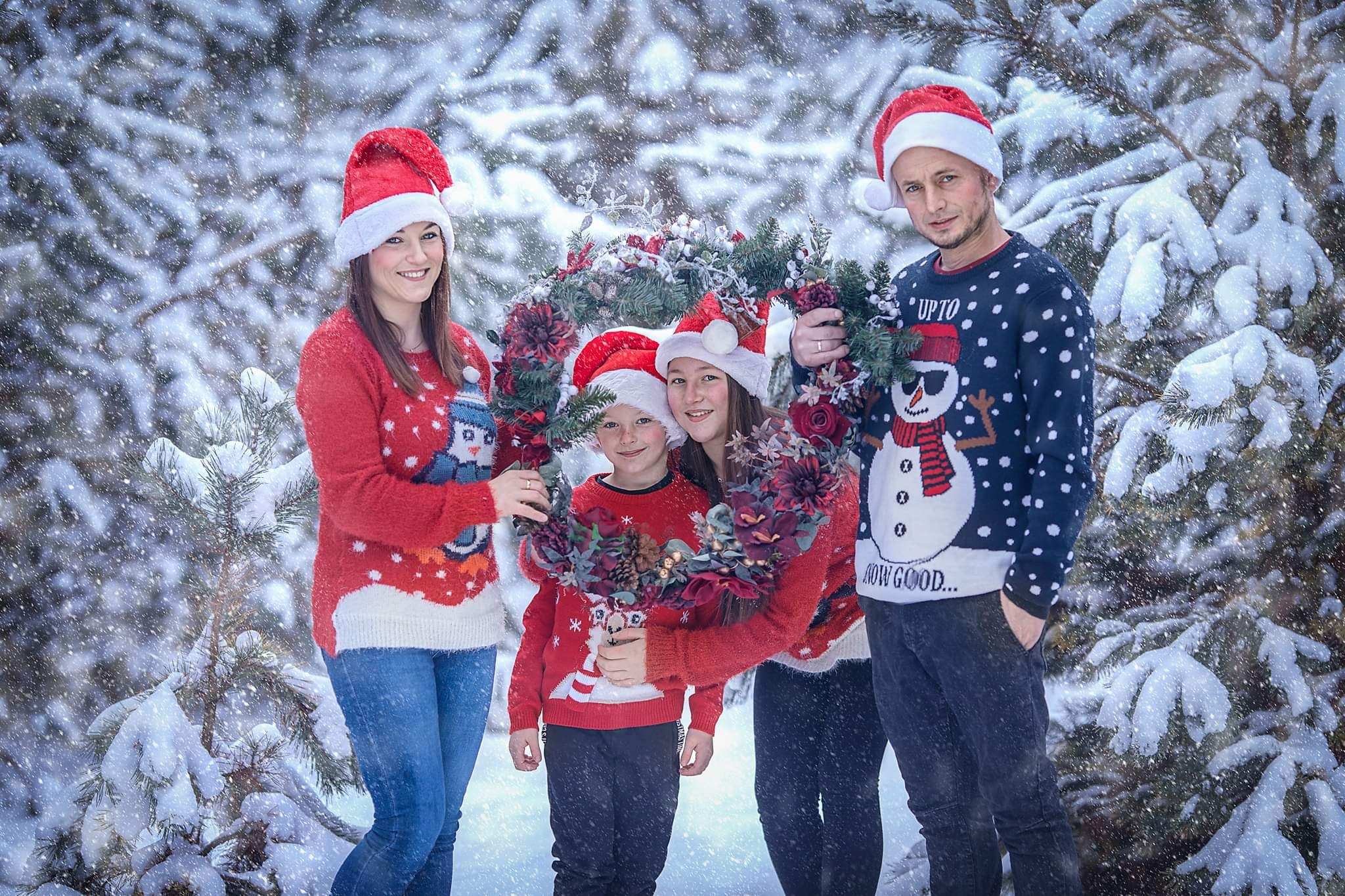 Uśmiechnięta rodzina w świątecznych swetrach i czapkach Mikołaja pozuje z wieńcem na tle zasypanego śniegiem lasu.
