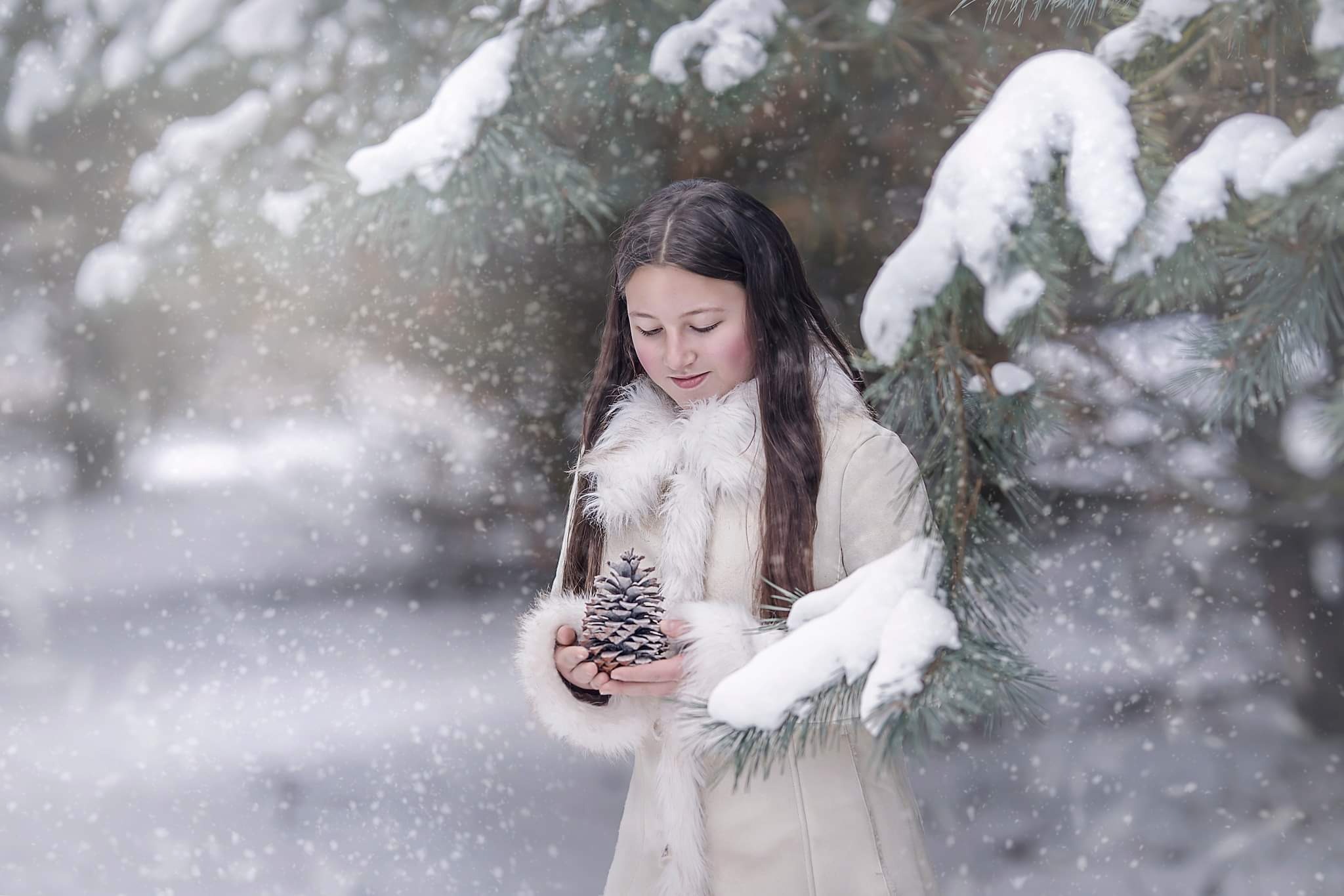 Nastolatka w białym płaszczu z futrzanym wykończeniem, trzymająca szyszkę na tle ośnieżonych gałęzi i padającego śniegu.