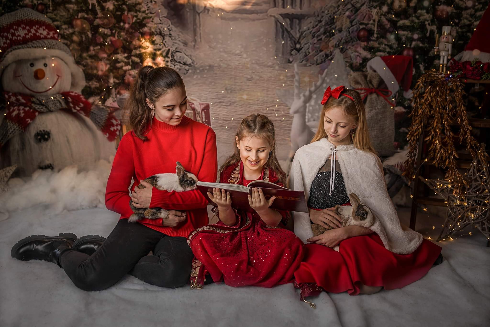 Trzy dziewczynki w świątecznych strojach siedzą na tle zimowej scenerii, czytając książkę i trzymając króliki.