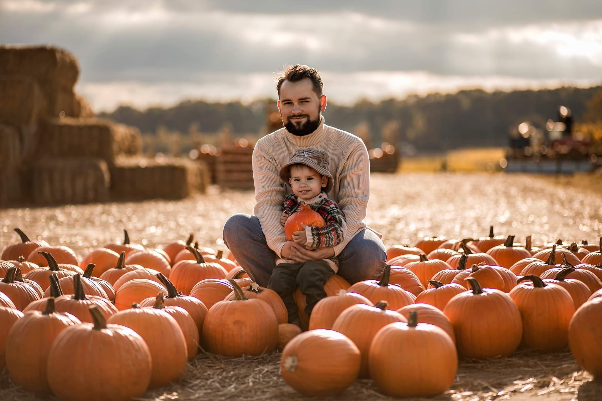Uśmiechnięty ojciec z synkiem siedzą w polu dyń, w tle stogi siana i drzewa.