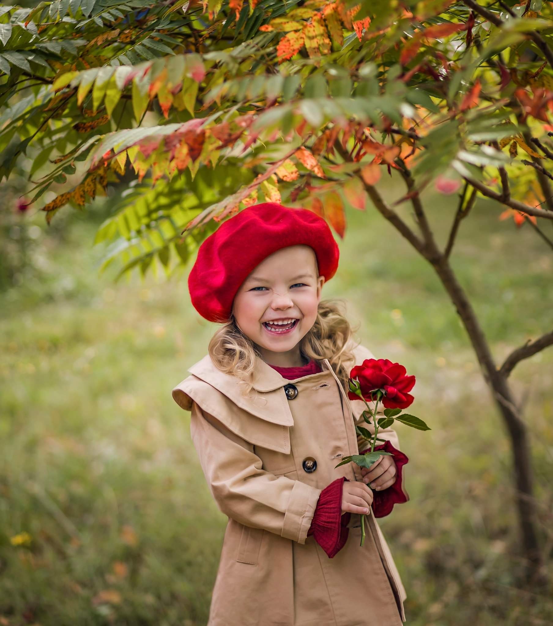 Uśmiechnięta dziewczynka w czerwonym berecie i beżowym płaszczu trzymająca czerwoną różę pod drzewem o jesiennych liściach.