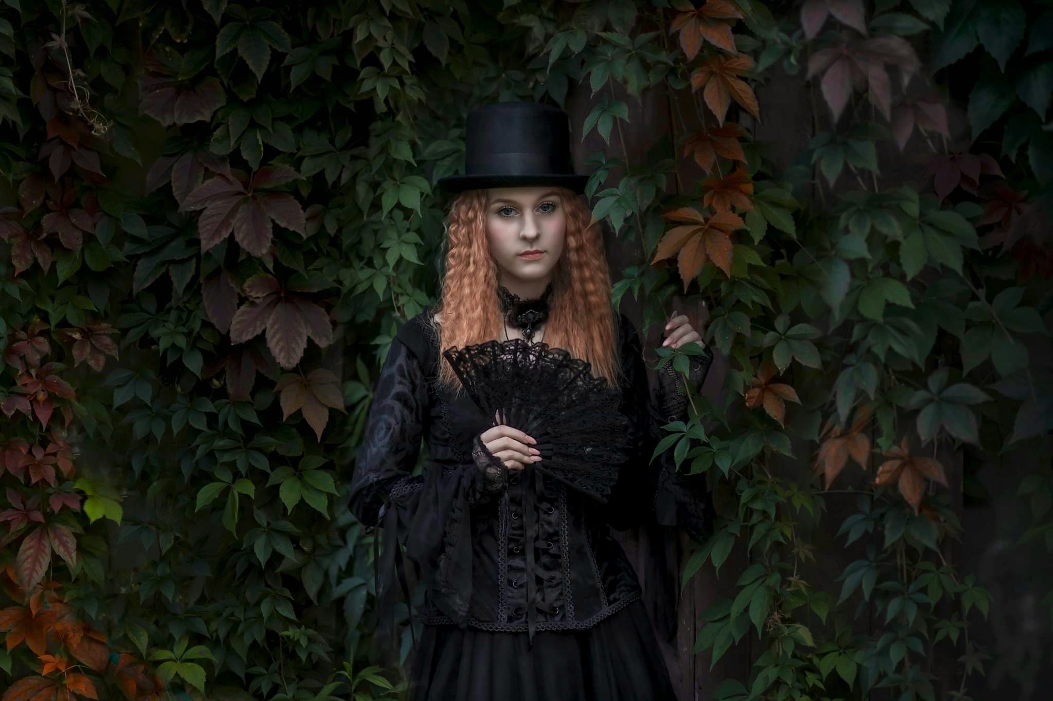 Portret kobiety w gotyckiej stylizacji z cylindrem i wachlarzem na tle ściany porośniętej bluszczem.