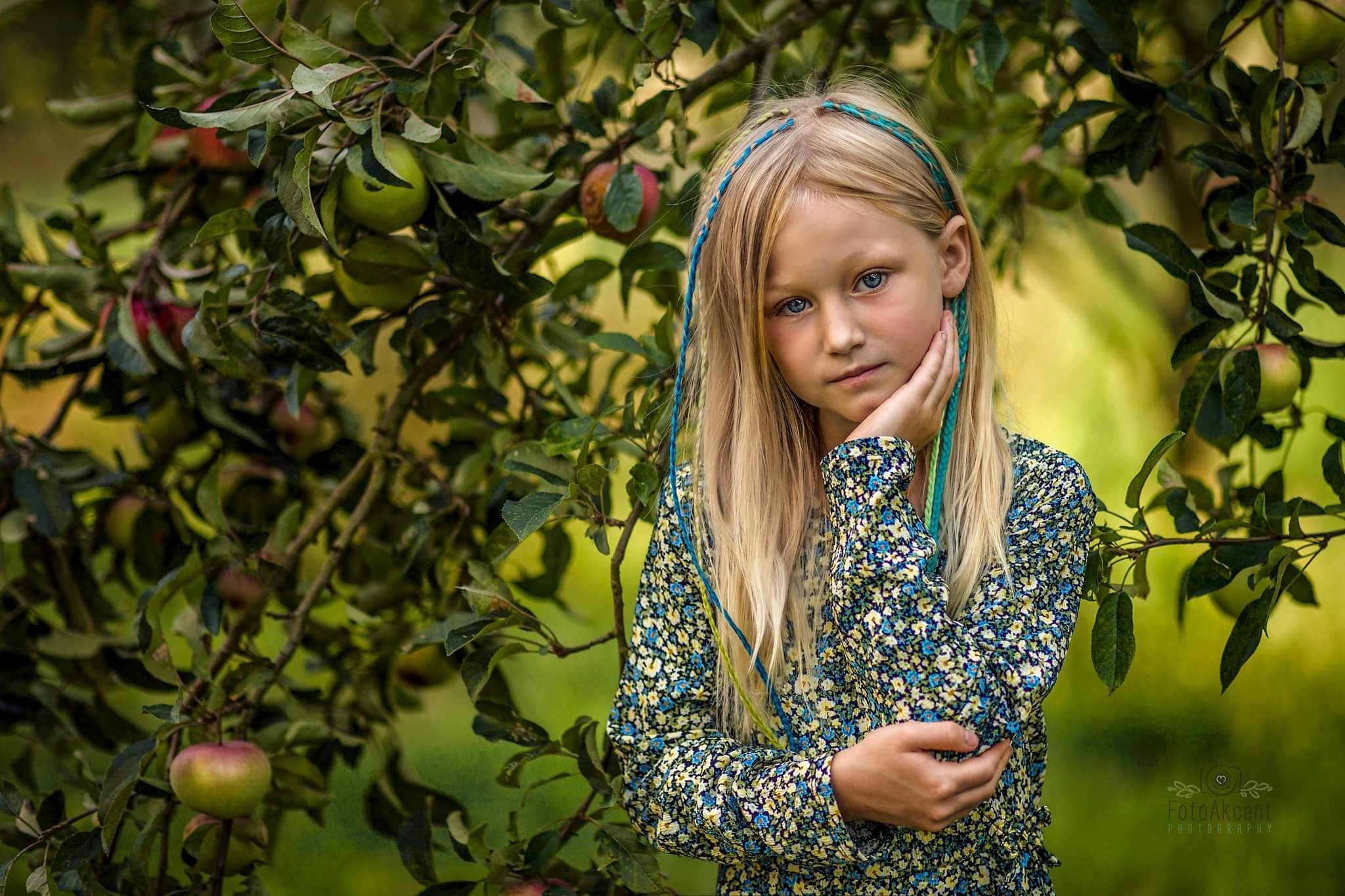 Portret blond dziewczynki z niebieskimi warkoczykami na tle jabłoni z dojrzałymi owocami. Ujęcie z bliska, miękkie światło.
