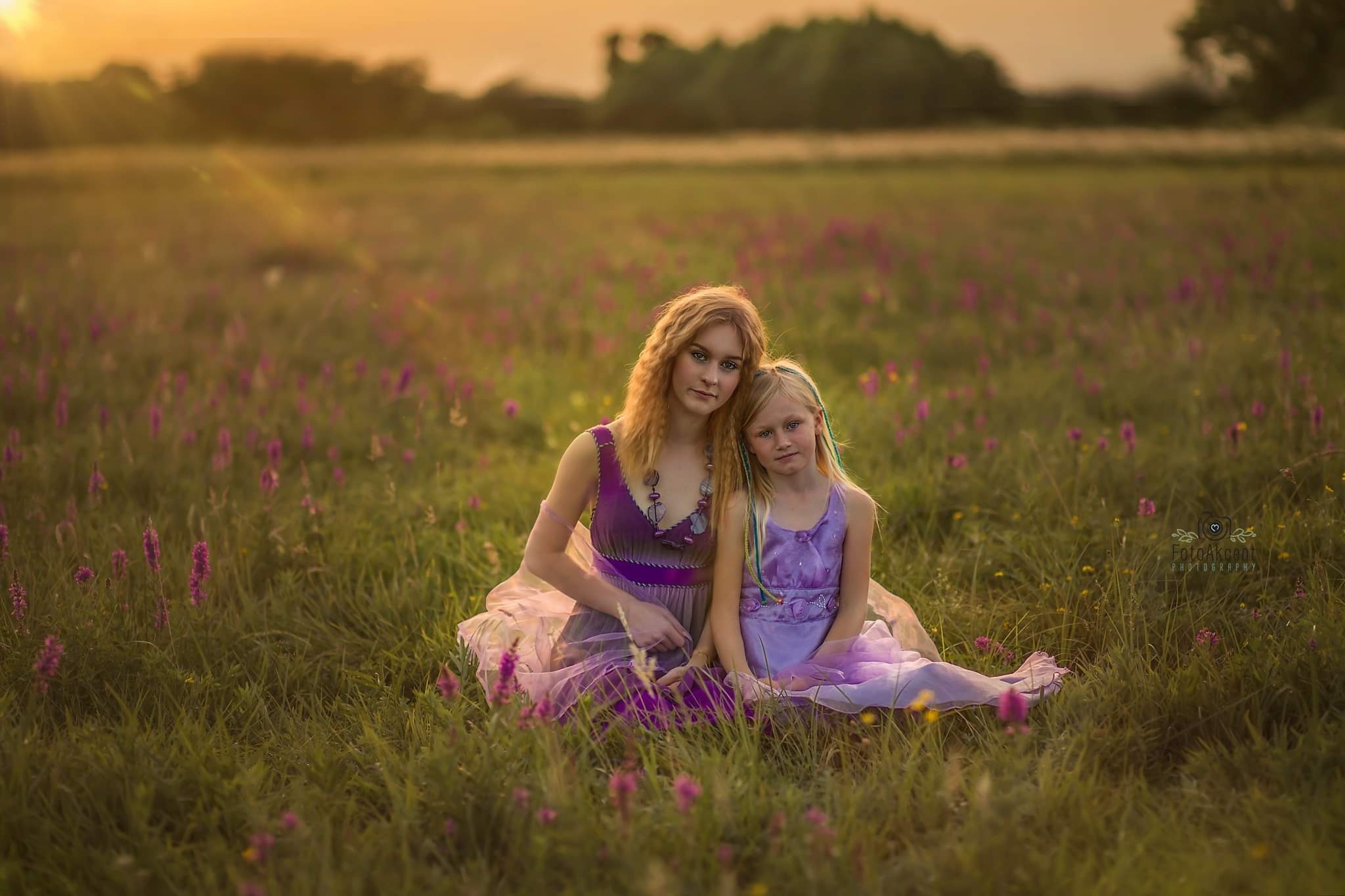 Dwie dziewczynki w fioletowych sukienkach siedzą na łące pełnej fioletowych kwiatów podczas zachodu słońca, z widocznym logo fotografa w prawym dolnym rogu.