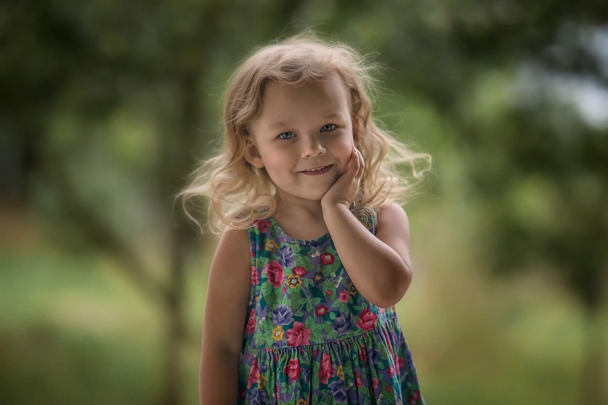 Portret uśmiechniętej dziewczynki z blond włosami, w kwiecistej sukience, pozującej na rozmytym zielonym tle.