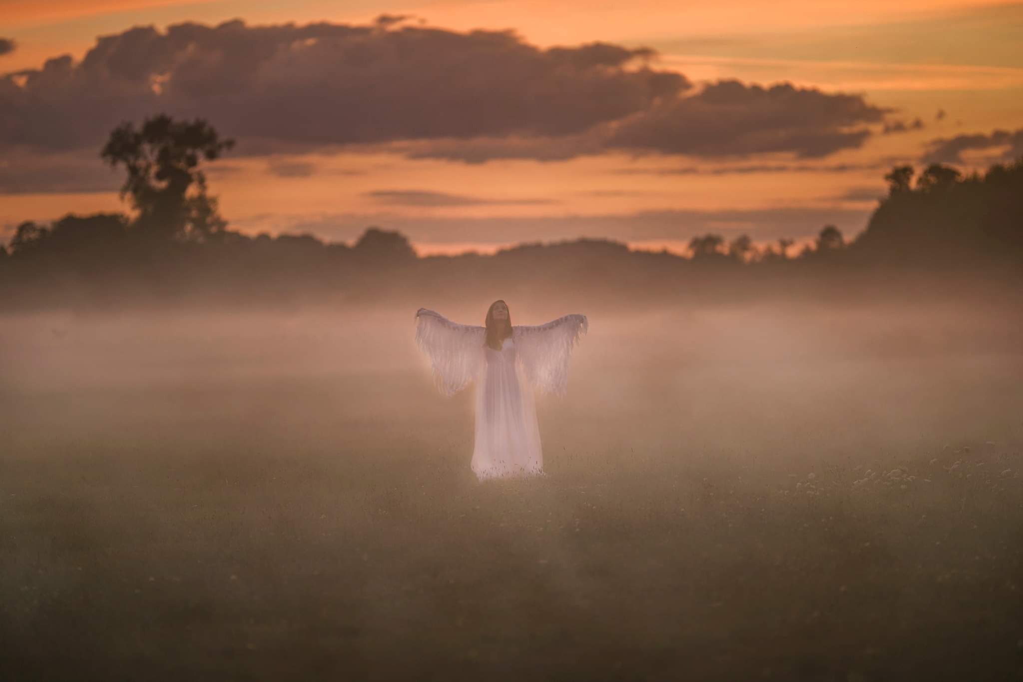 Kobieta w białej sukni z rozpostartymi ramionami, otulona mgłą na tle zachodzącego słońca, tworząca eteryczny portret w plenerze.