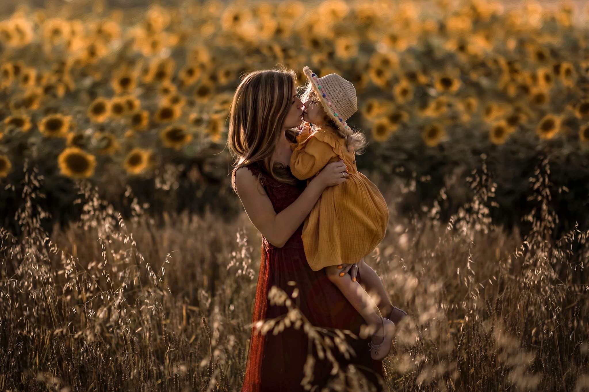 Matka w bordowej sukience trzyma córeczkę w żółtej sukience i kapeluszu na tle pola słoneczników, delikatny pocałunek.
