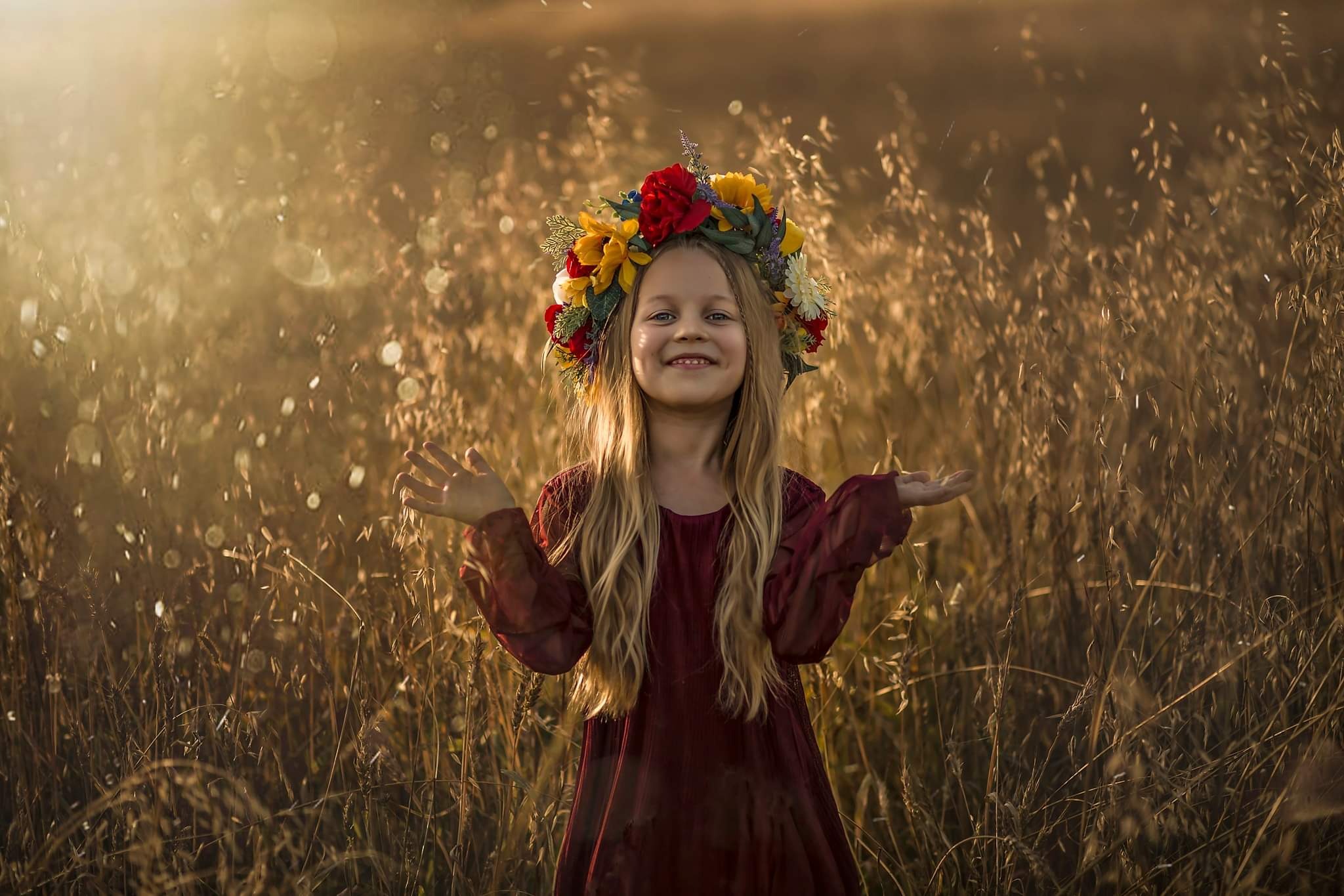 Uśmiechnięta dziewczynka z blond włosami i wiankiem z kolorowych kwiatów na głowie, stojąca na tle wysokiej trawy w złocistym świetle zachodzącego słońca, z rozłożonymi rękami.