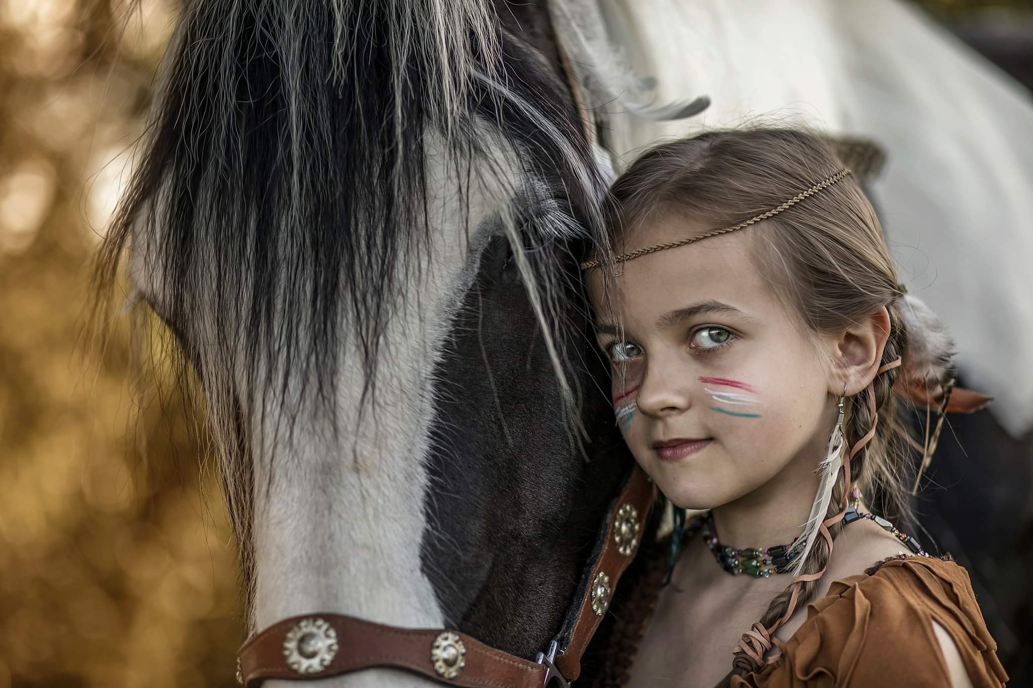 Dziewczynka z pomalowaną twarzą w stylu indiańskim, oparta o pysk konia z bogato zdobioną uprzężą, z piórami we włosach i naszyjnikiem z koralików, w miękkim, naturalnym oświetleniu.