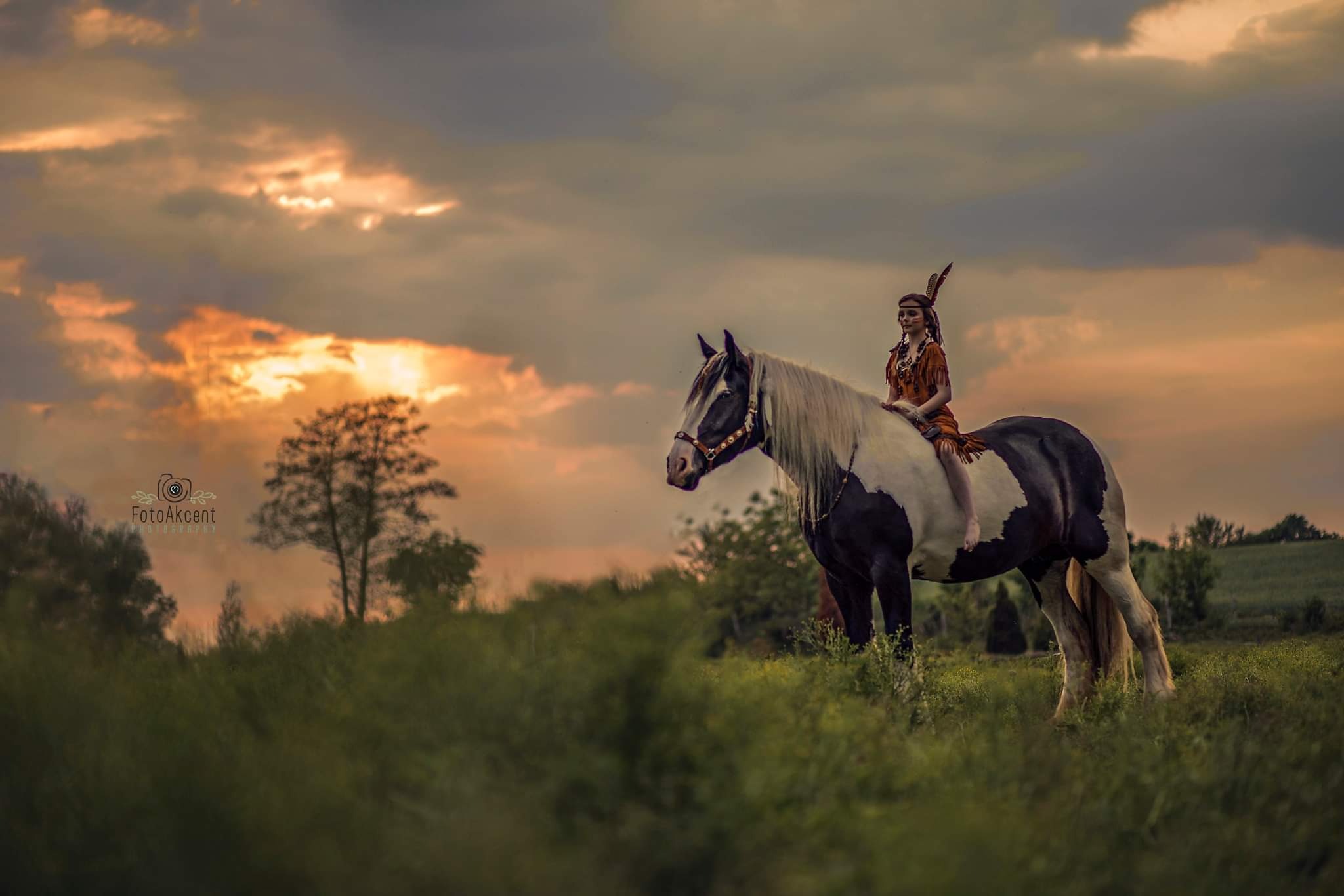 Kobieta w stroju inspirowanym kulturą rdzennych Amerykanów siedzi okrakiem na koniu rasy Pinto w trawiastym terenie o zmierzchu, z pomarańczowo-szarym niebem w tle.