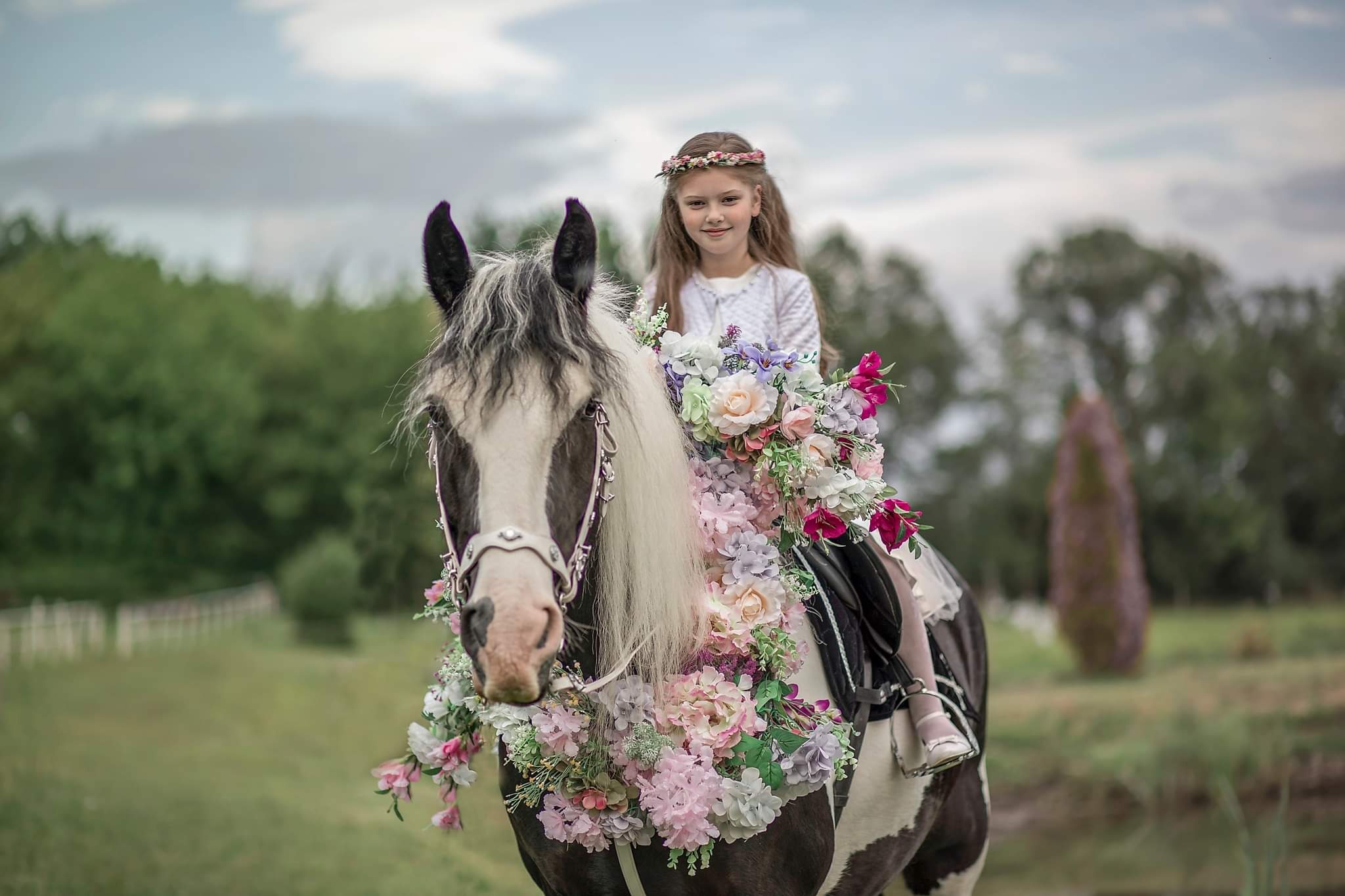Dziewczynka w wianku na głowie siedzi na koniu udekorowanym kwiatami w plenerze.