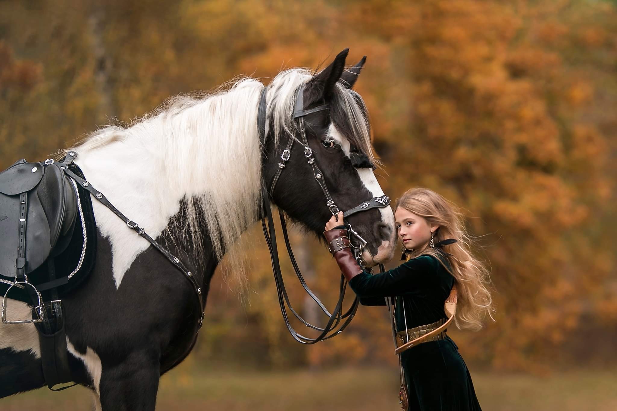 Dziewczynka w zielonej sukni, z długimi blond włosami, trzyma czarno-białego konia za uzdę na tle jesiennych drzew.