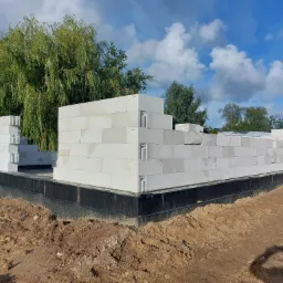 Budowa domu murowanego z bloczków w Pucku, widoczna czarna izolacja fundamentów i częściowo wymurowane ściany parteru na tle drzew i błękitnego nieba.