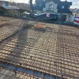 Zbrojenie płyty fundamentowej przygotowane do zalania betonem, widoczny betonowóz z pompą w tle, drewniane szalunki i plastikowe dystanse.