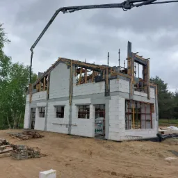 Surowy stan domu murowanego z betonu komórkowego, widoczny dźwig podający beton, drewniane szalunki okienne, teren budowy z materiałami budowlanymi i drzewami w tle.