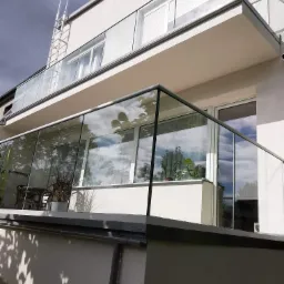 Dwukondygnacyjny budynek z balkonami zabezpieczonymi szklanymi balustradami z szarymi ramami, na górnym balkonie widoczna drabina aluminiowa.