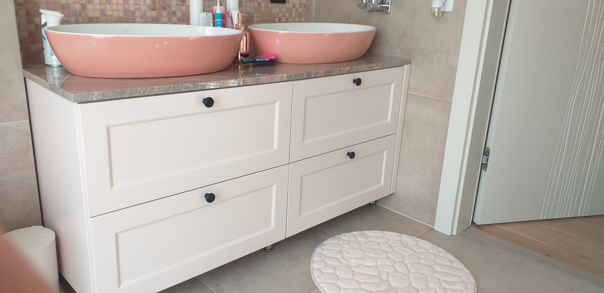 Jasna szafka łazienkowa z dwoma umywalkami w kolorze łososiowym, blat z granitu, okrągły dywanik, minimalistyczny design.