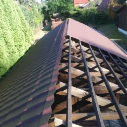 Widok z góry na konstrukcję dachu z częściowo położonym, bordowym pokryciem blachodachówką falistą, widoczna więźba dachowa i okolica z zielenią i zabudowaniami.