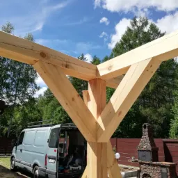 Surowa konstrukcja drewniana, prawdopodobnie altany, w trakcie budowy w Nidzicy, z widocznym samochodem dostawczym i kamiennym grillem w tle.