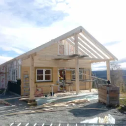 Wznoszenie szkieletu drewnianego domu jednorodzinnego na fundamencie, widoczne elementy konstrukcyjne dachu, ściany pokryte płytami, piła stołowa na rusztowaniu, teren budowy z widokiem na wzgórza.