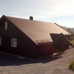 Ciemny, parterowy budynek z dwuspadowym dachem pokrytym brązowym gontem bitumicznym, z małym oknem na szczycie i kominem, otoczony żwirowym podłożem i zielenią.