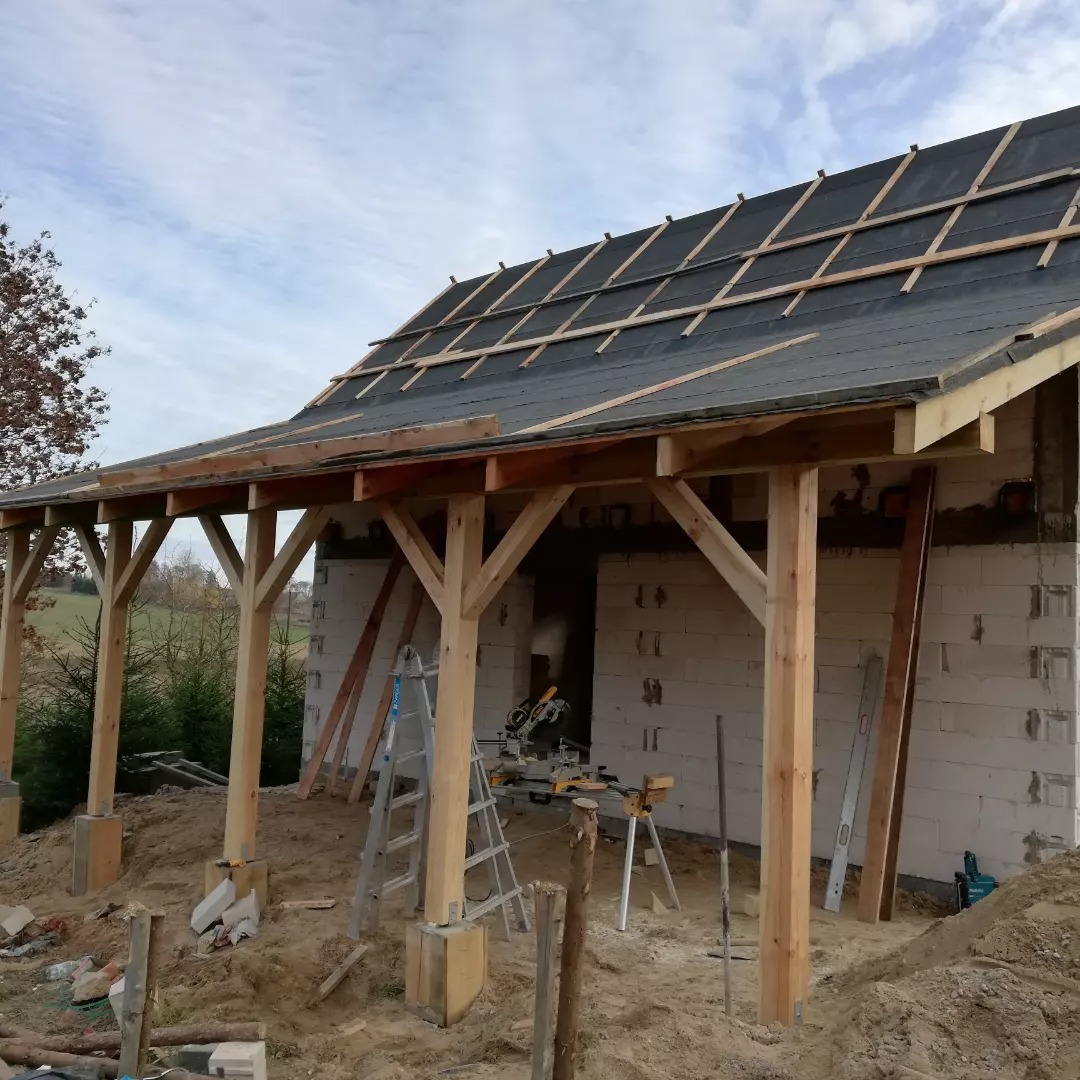 Budowa zadaszenia z drewnianymi słupami i konstrukcją dachu pokrytą papą, widoczna piła stołowa na statywie i drabina aluminiowa, tło stanowi krajobraz polny.