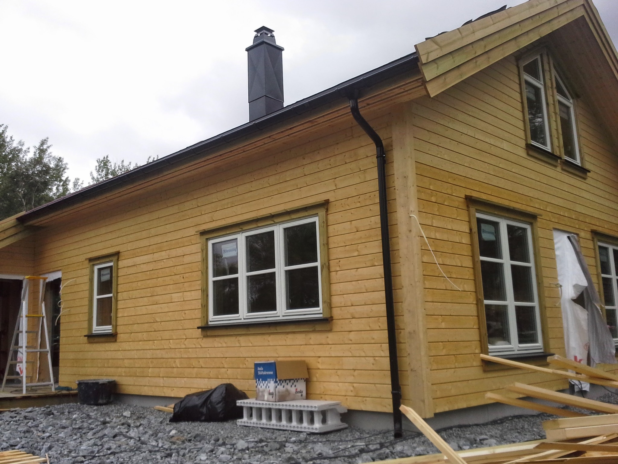 Surowy dom szkieletowy z jasnego drewna w trakcie budowy, widoczne okna z białymi ramami, czarny komin, rynna, drabina i materiały budowlane na żwirowym podłożu.