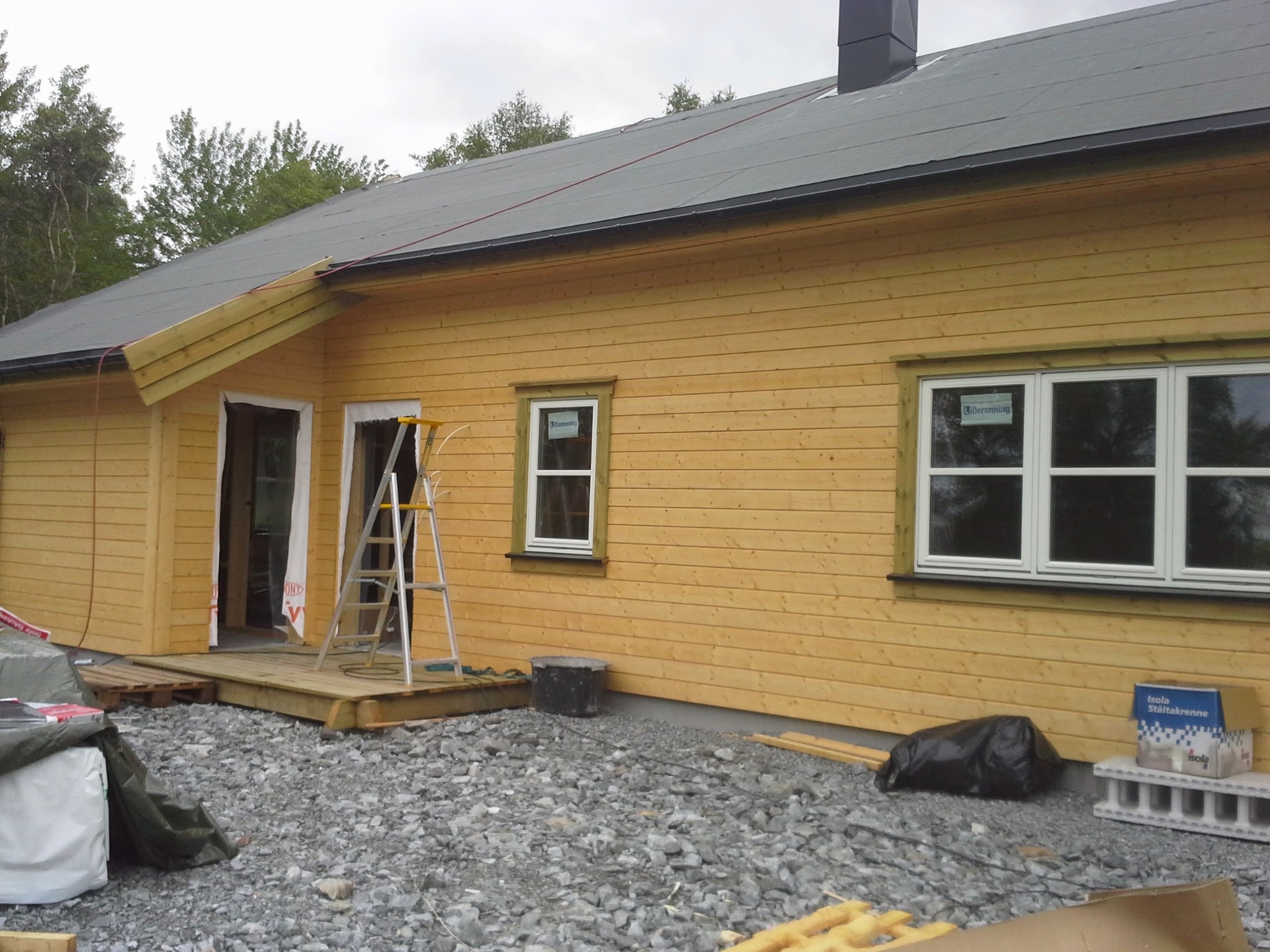 Żółty dom w trakcie budowy z widoczną konstrukcją dachu, oknami z naklejkami, schodami i gruzem na zewnątrz.