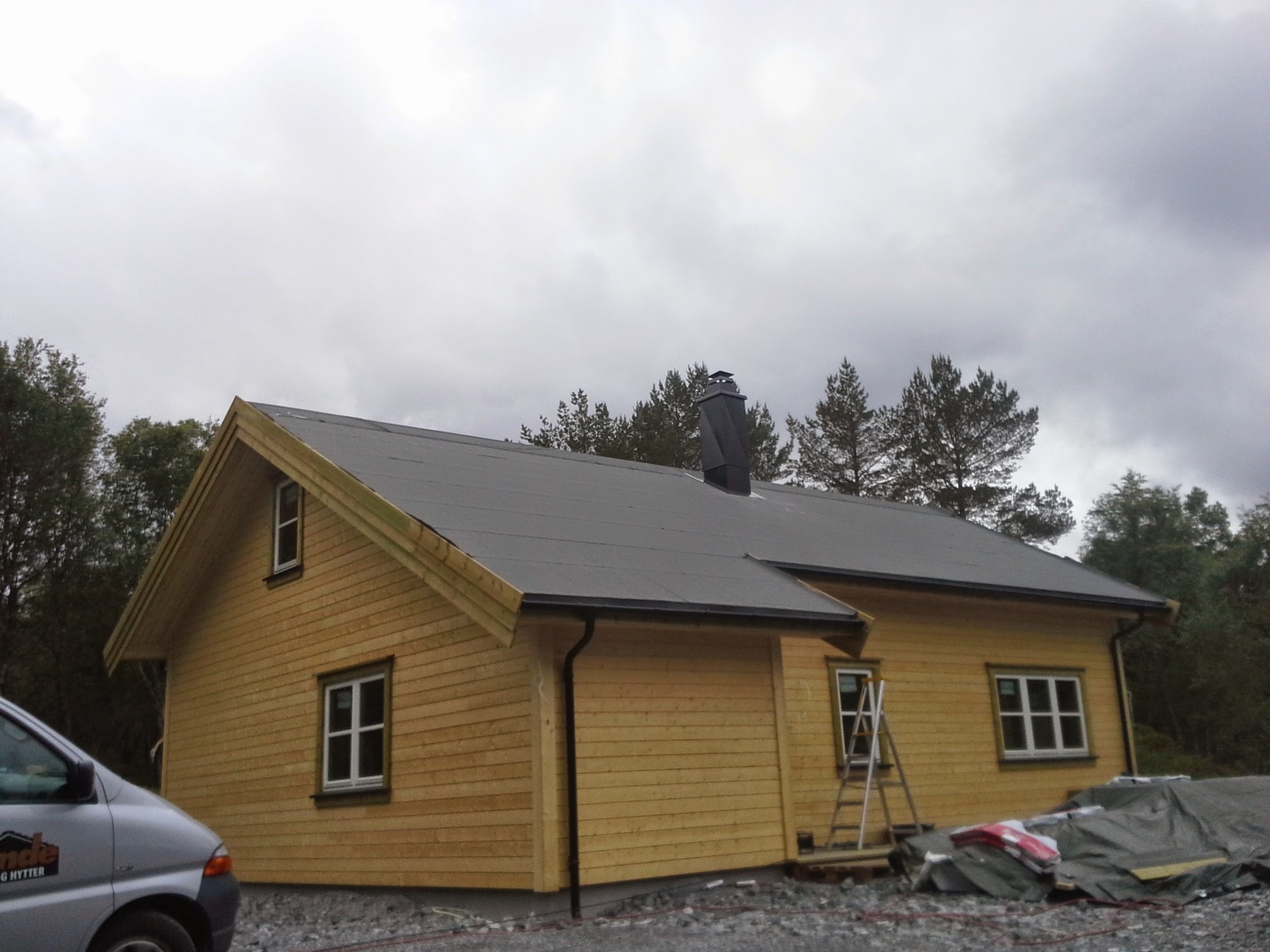 Żółty dom w trakcie budowy z szarym dachem i czarnym kominem, drabina oparta o ścianę, widoczny samochód dostawczy firmy.