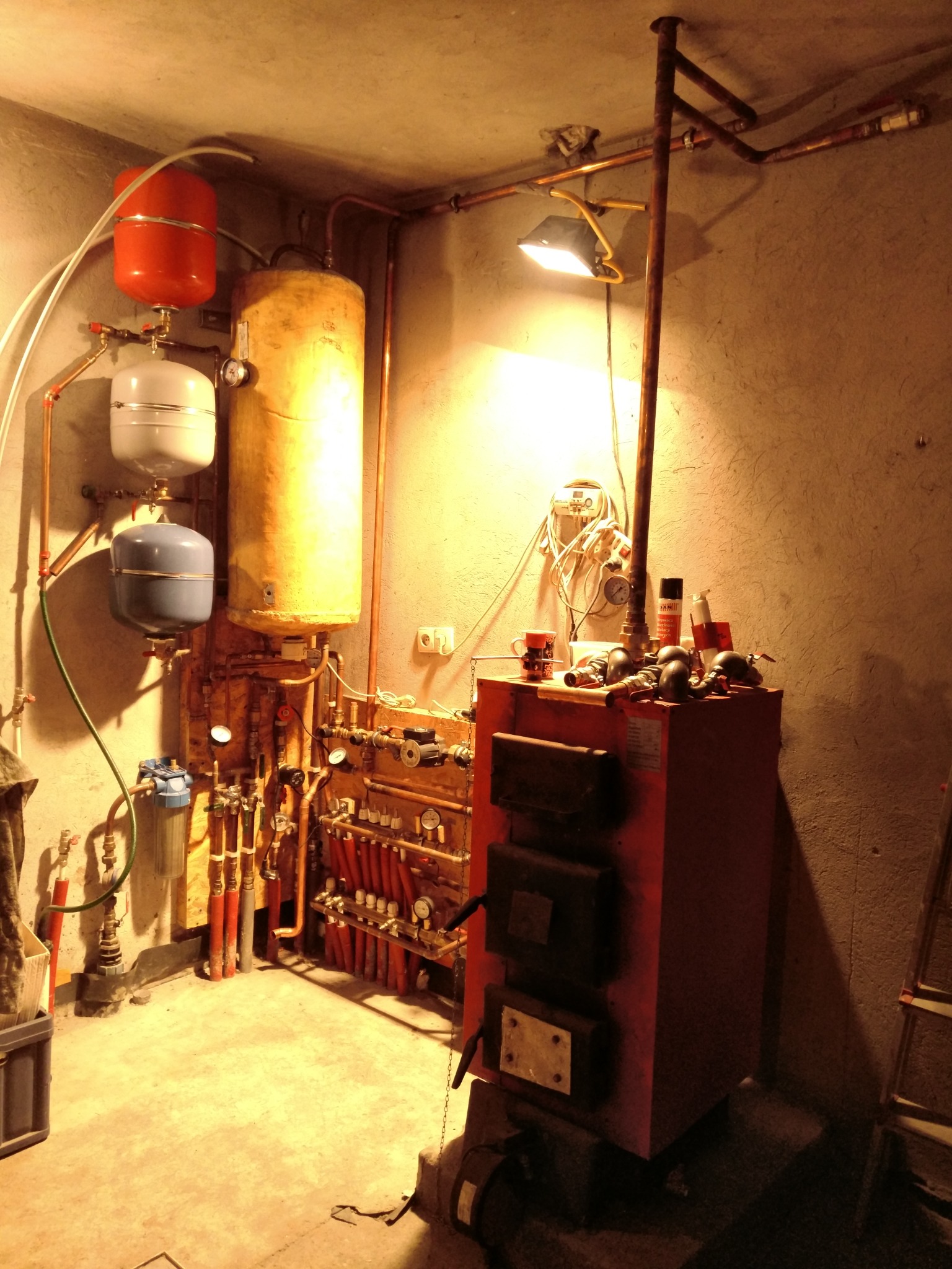 Instalacja centralnego ogrzewania z piecem na paliwo stałe, zbiornikami wyrównawczymi i rozdzielaczem, oświetlona lampą halogenową w piwnicy.