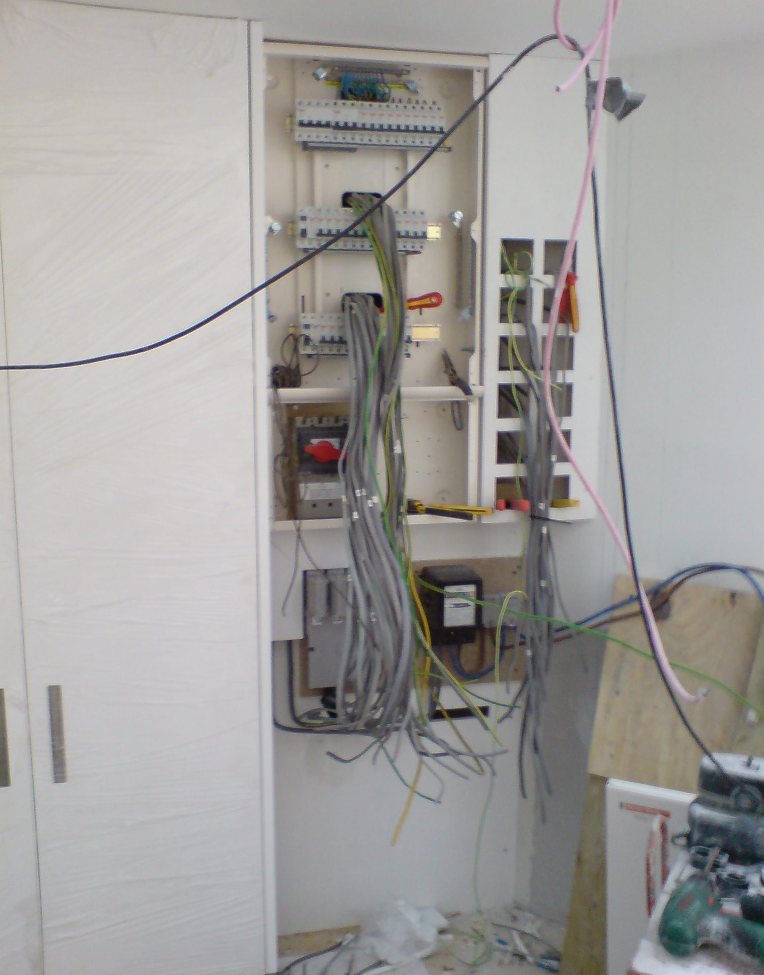 Wnętrze szafy z rozdzielnicą elektryczną w trakcie montażu instalacji. Widoczne liczne przewody, bezpieczniki, narzędzia i elementy montażowe. Na pierwszym planie wiertarka i płyta OSB.